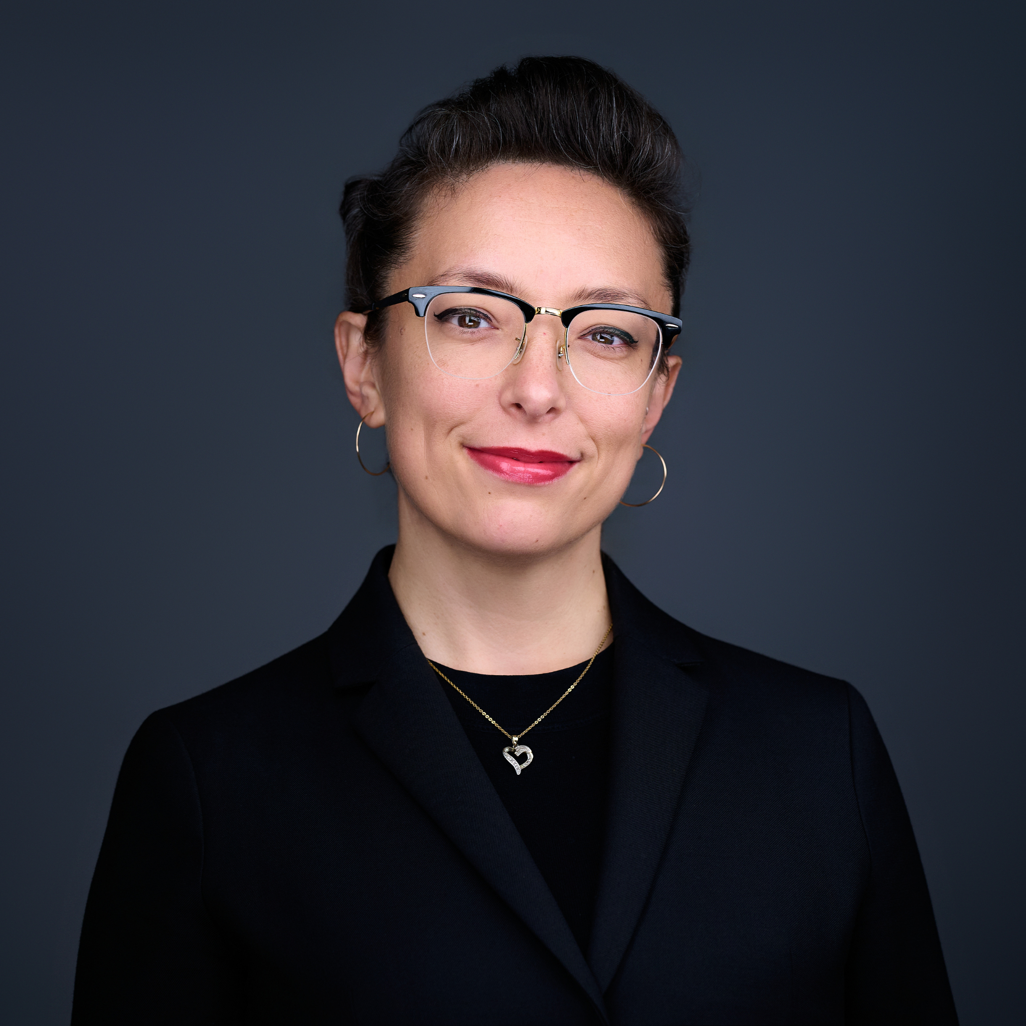 Kim Roney square Kim Romney, a white woman with short hair, glasses, and red lipstick, in front of a grey background.